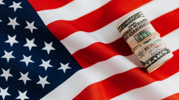 Foto colorida horizontal. Cédulas de dinheiro enroladas com elástico sobre uma bandeira dos Estados Unidos.
