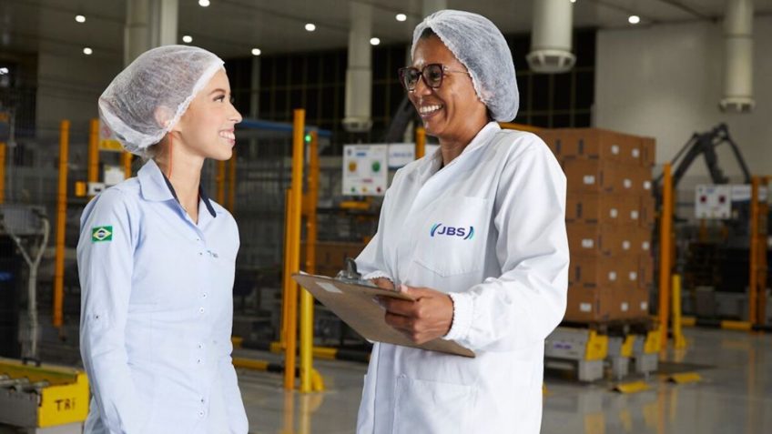 Duas funcionárias da JBS sorriem uma para a outra. Ambas usam jaleco branco e touca branca. Estão em uma área industrial