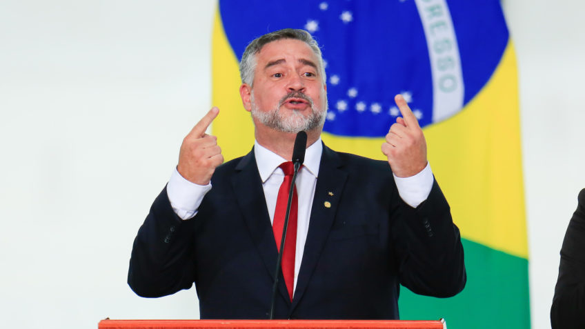 Paulo Pimenta é um homem branco, grisalho. Usa terno preto, camisa branca e gravata vermelho. Fala em um microfone. No fundo, há uma bandeira do Brasil.